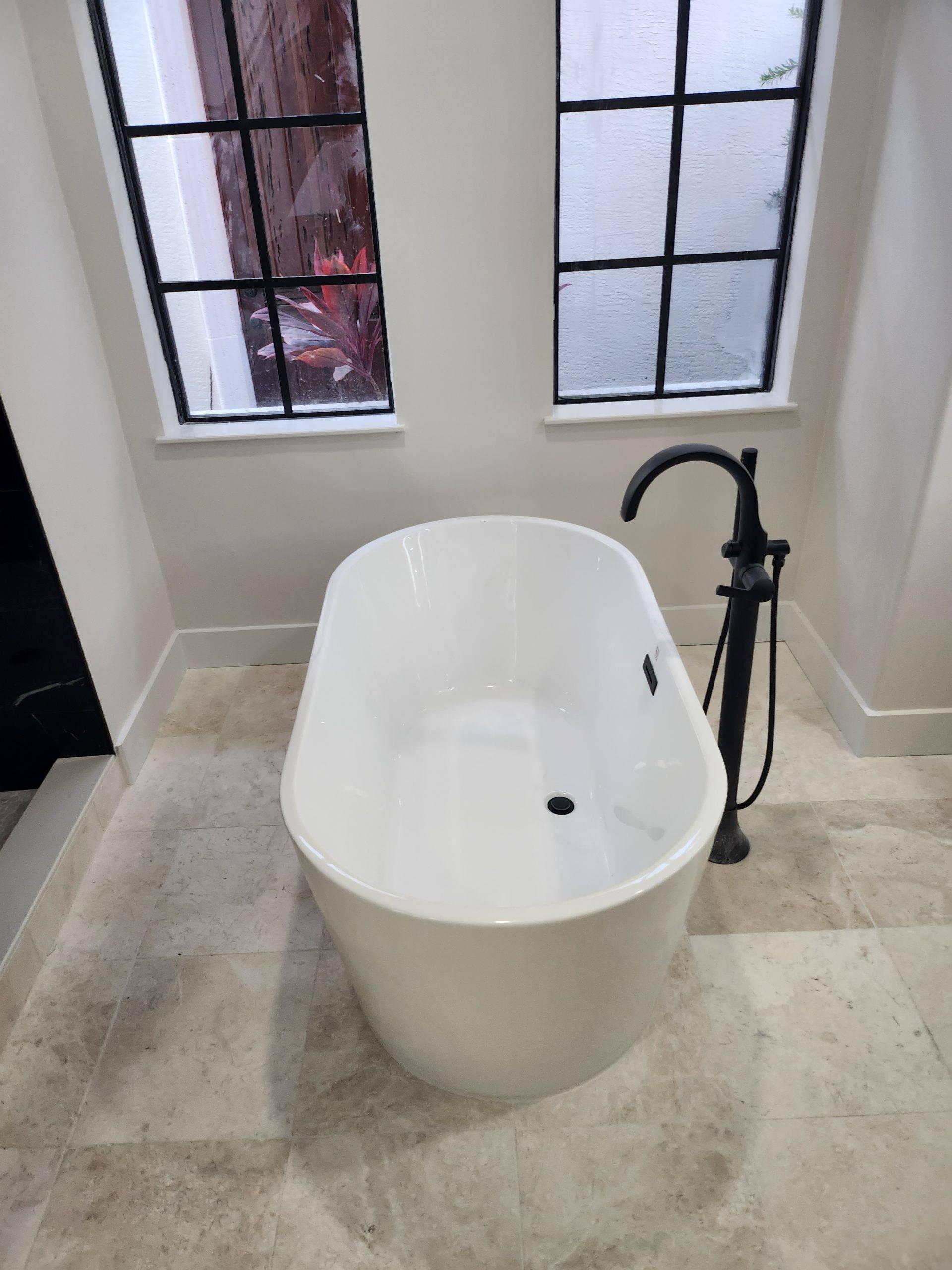 Freestanding white soaking tub with a matte black floor-mounted faucet and a travertine-look tile floor