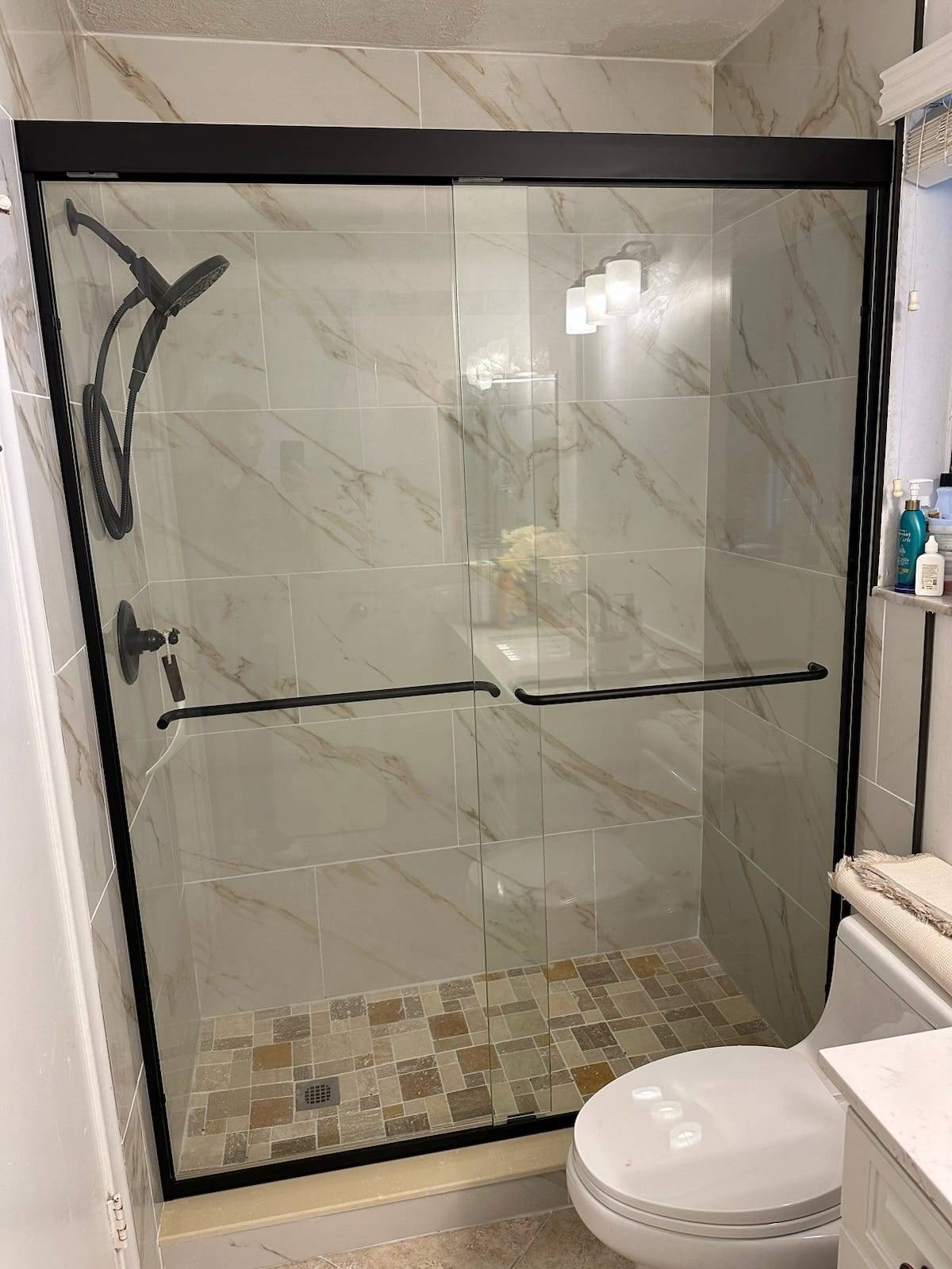 Tiled shower remodel with marble-look tile, black-framed sliding glass doors, and a mosaic shower floor